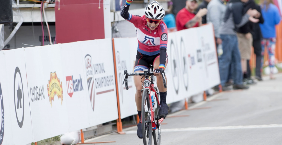 Erica Clevenger, on her bicycle wearing a UA jersey, raises her fist above her head in victory