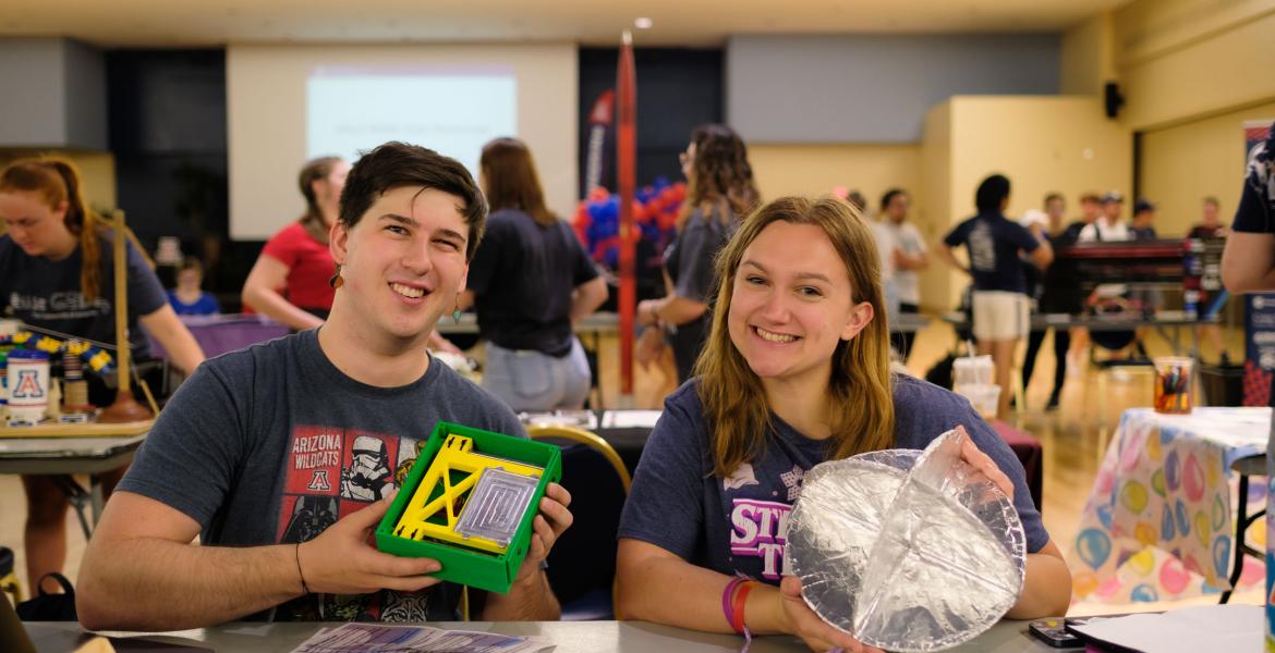 Two students sit at a booth for the Engineering Club showcase and smile. He is holding a green and yellow box and she is holding a circular metallic object..