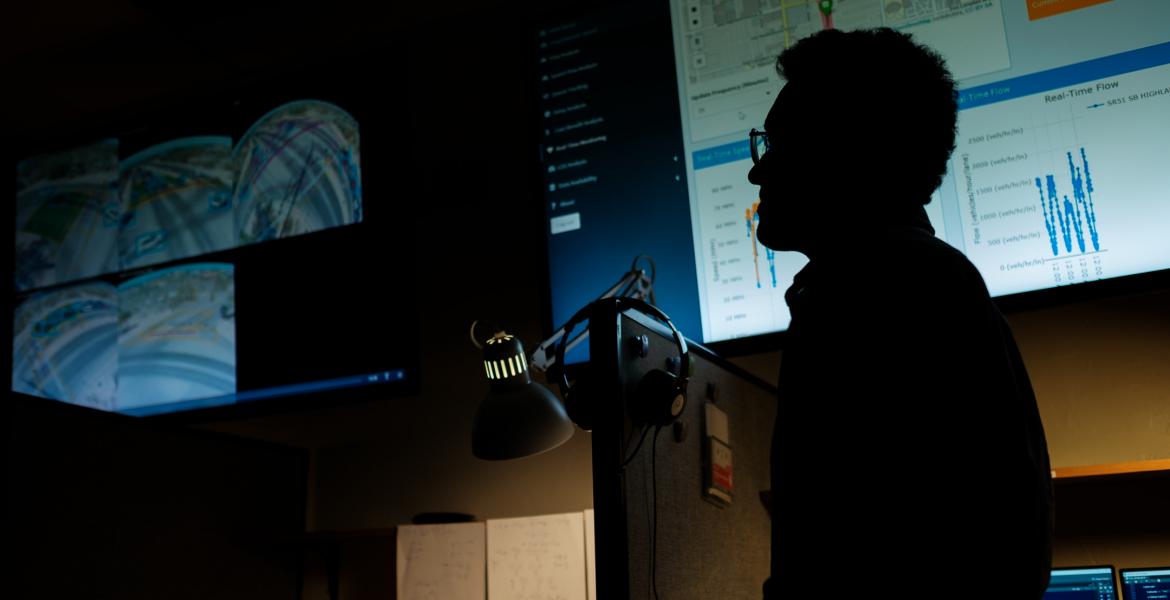 A silhouette of a man wearing glasses in front of a large computer screen.
