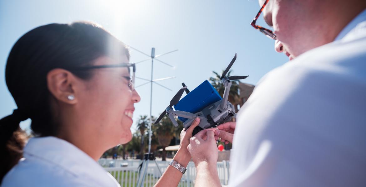 two students, seen from behind, examine a drone. the sun is shining and they are smiling.