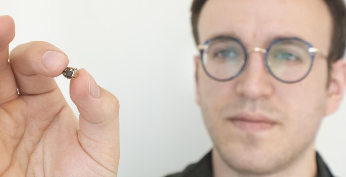 A man holds up a tiny electronic device, about the size of a chocolate chip.