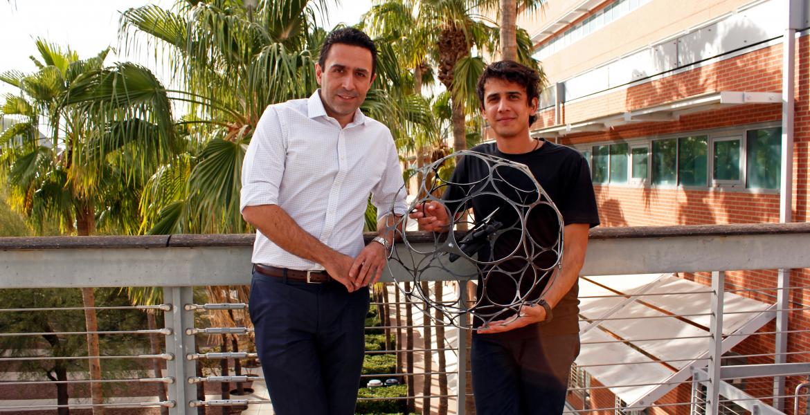 Two men stand in front of a railing and some palm trees, holding a drone encased in a spherical cage, about three feet in diameter