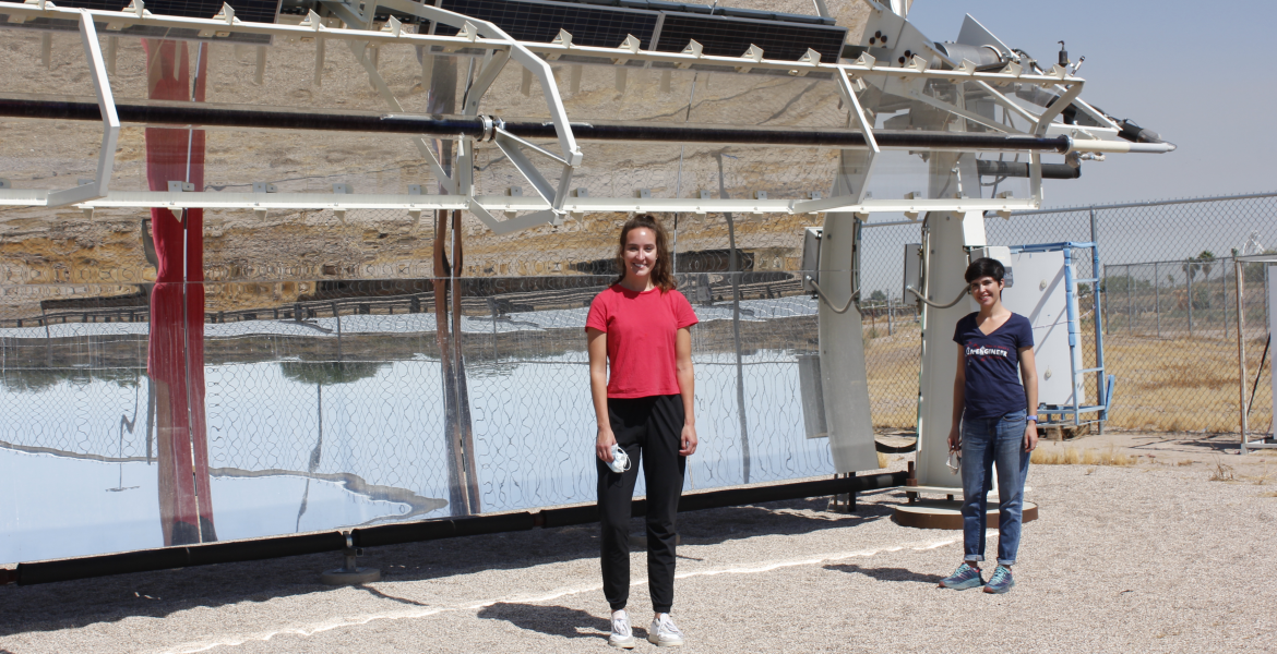 Two women stand in front of a solar-powered desalination system, shaped like a giant curved mirror