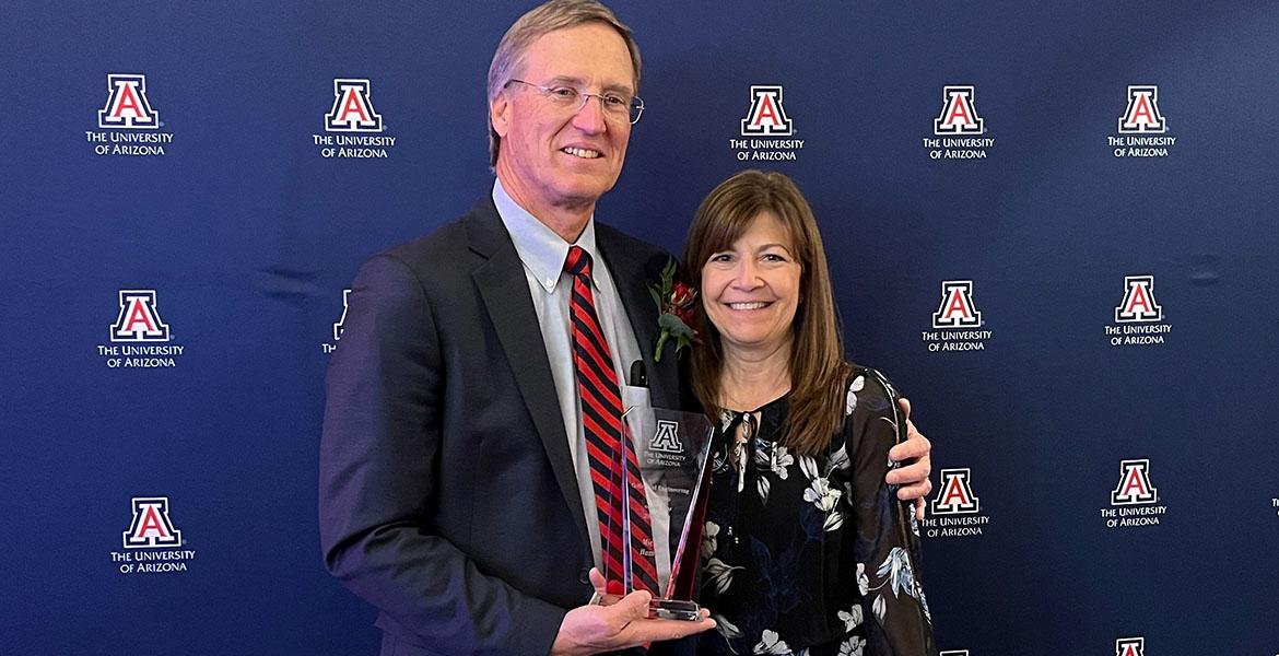 Mike and Sheri Hummel in front of a UA backdrop with a glass award