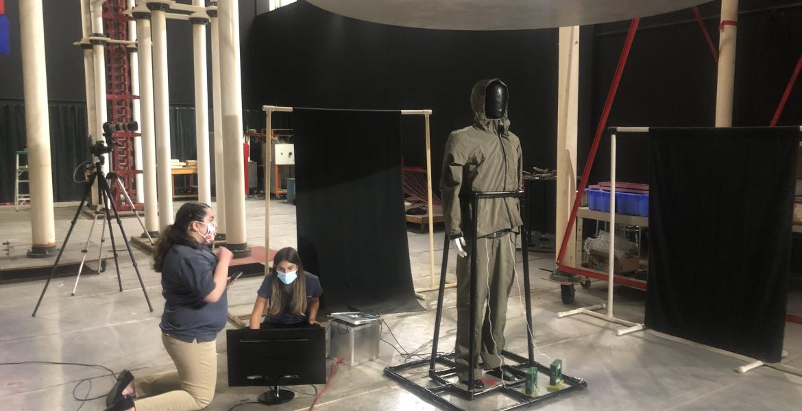 A mannequin covered in black tape is held up by a PVC pipe frame in a large room. Two students kneel next to electronic equipment near the mannequin.