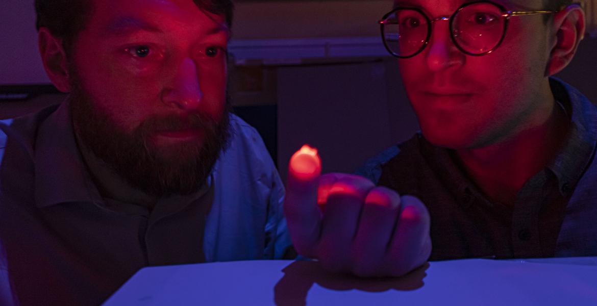 Two men, visible from the shoulders up, in a dimly lit room. The man on the left is holding a small, reddish light source on his hand, which is illuminating their faces. Both are staring at the light.