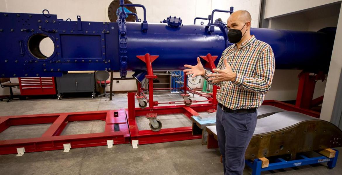 Jesse Little stands in front of a large blue wind tunnel -- it looks like a horizontal pipe -- on red scaffolding. He is wearing a black mask over his nose and mouth and gesturing with his hands to explain something.