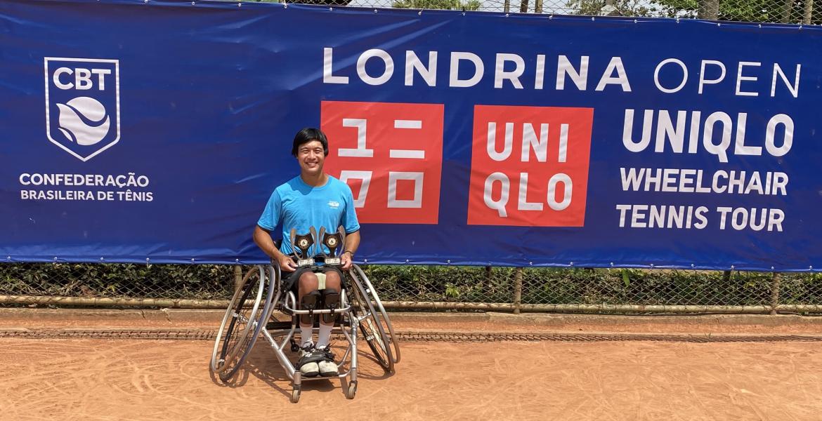 Jason Keatseangsilp sits in a sports wheelchair with slanted wheels, smiling and holding two trophies on his lap. Behind him is a blue banner that reads, 