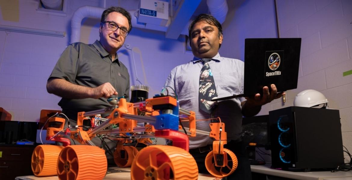 In the foreground, a robot with four orange wheels. Two men stand behind the robot.