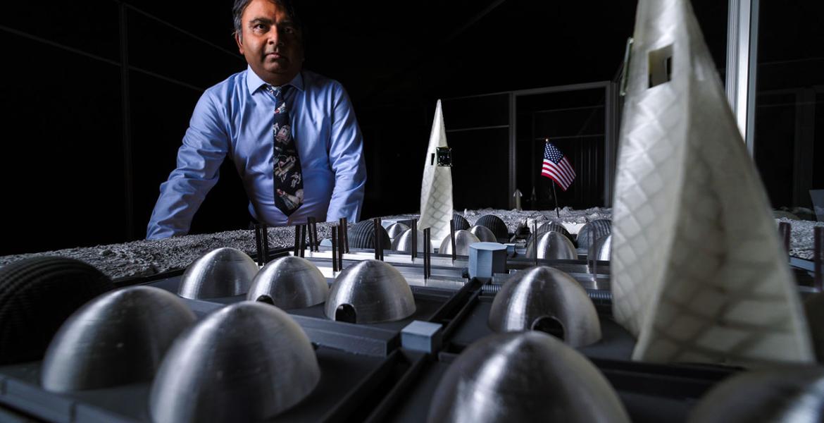 A man in a blue shirt stands in front of a small-scale table that mimics a lunar base.