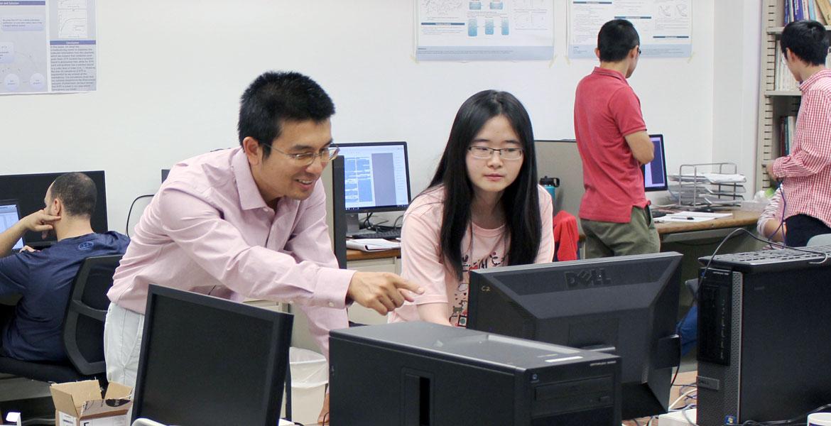 Ming Li leans over a computer, smiling and helping a student.