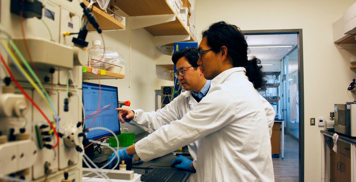 Two researchers in white lab coats look at a computer screen.