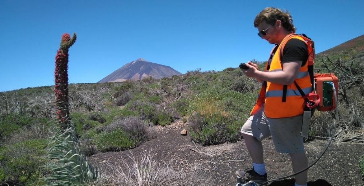 A young man in an orange vests stands in an area with volcanic rock.