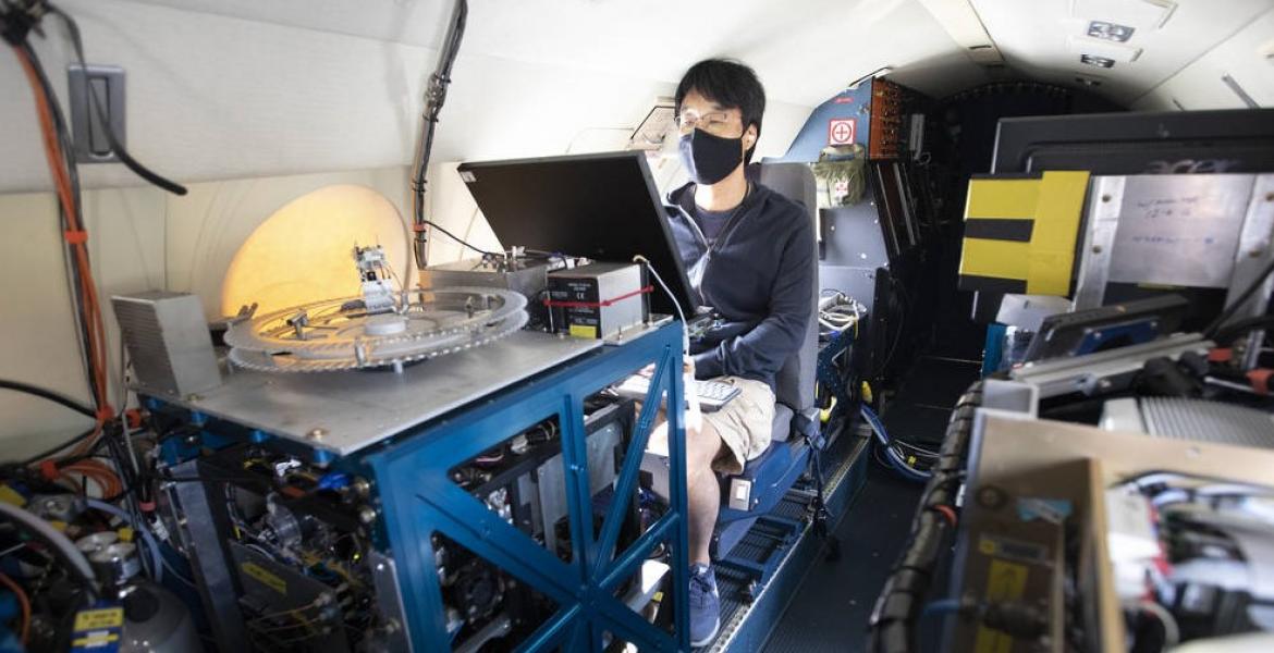 A young man wearing a face mask sits behind a monitor, surrounded by technology on a plane.