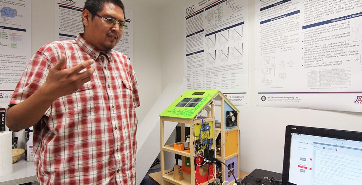 A student stands in front of a house model outfitted with electronic sensors and hooked to a monitor