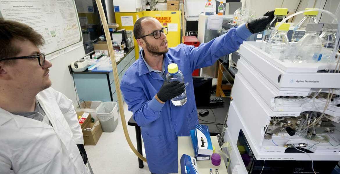 Two men in a laboratory reach for water samples setting on top of a machine.