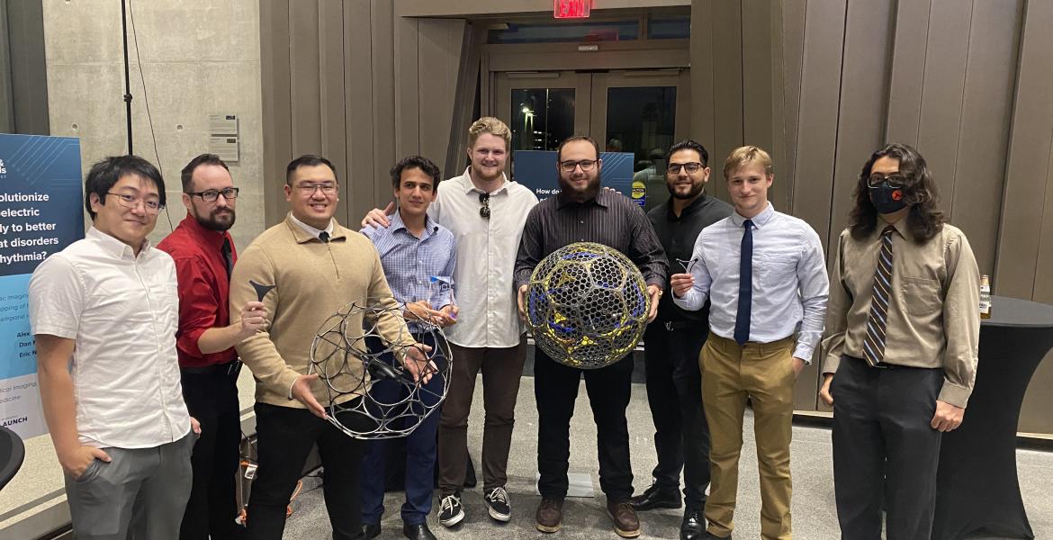 Nine men stand in a row. Two are holding large, spherical mesh robots.