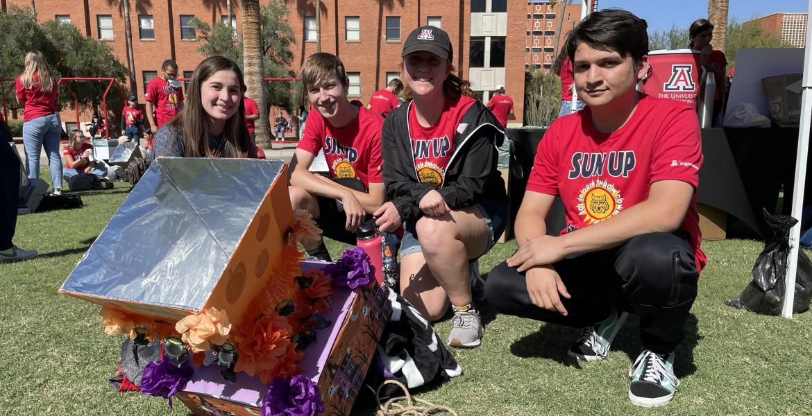 Four students in matching red t-shirts pose on the lawn next to their solar oven -- a cardboard box with a funnel shape on top, decorated with orange and purple tissue paper.