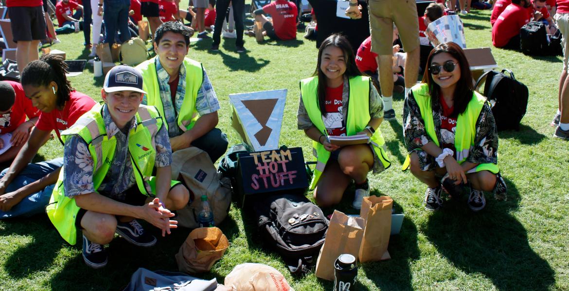 Two young men and two young women wearing Hawaiian shirts and bright yellow vests kneel for a photo next to their solar oven, a cardboard and aluminum foil device labeled with their group name: 