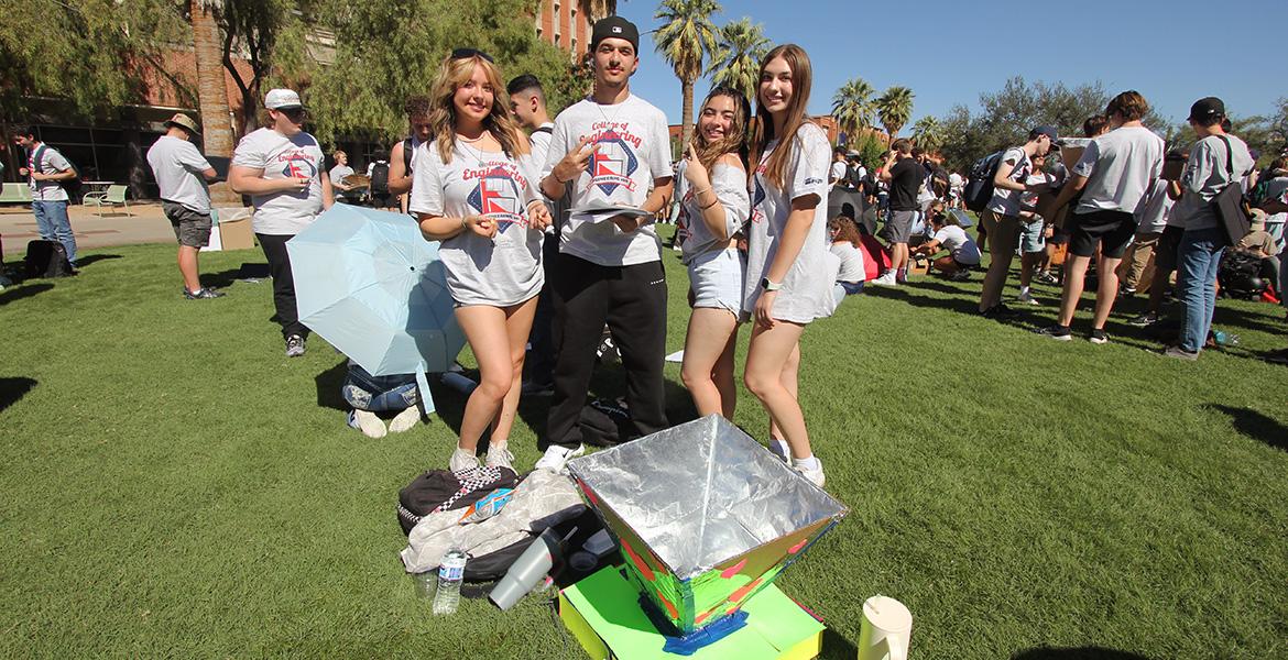 Four students in white shirts smile and pose for a photo on a grassy lawn with an oven made of aluminum foil and cardboard at their feet.