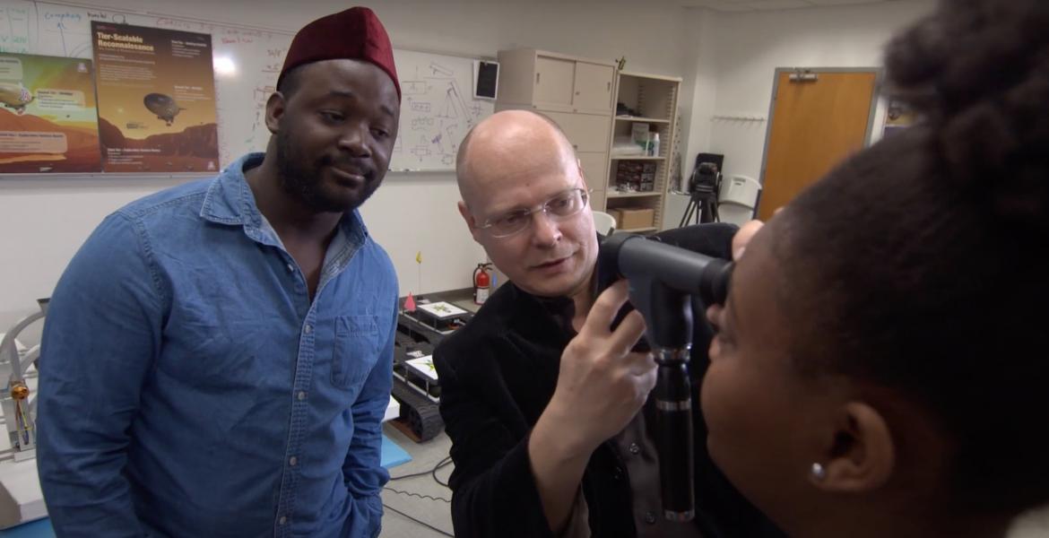 Wolfgang Fink uses an optical device to examine a young woman's eyes, while another young man looks on in the background.