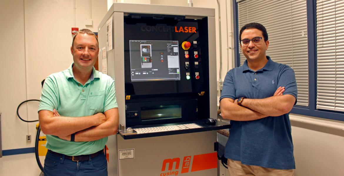 Andrew Wessman and Mohammed Shafae standing next to a 3D printer, which is about two feet wide and a bit taller than them.