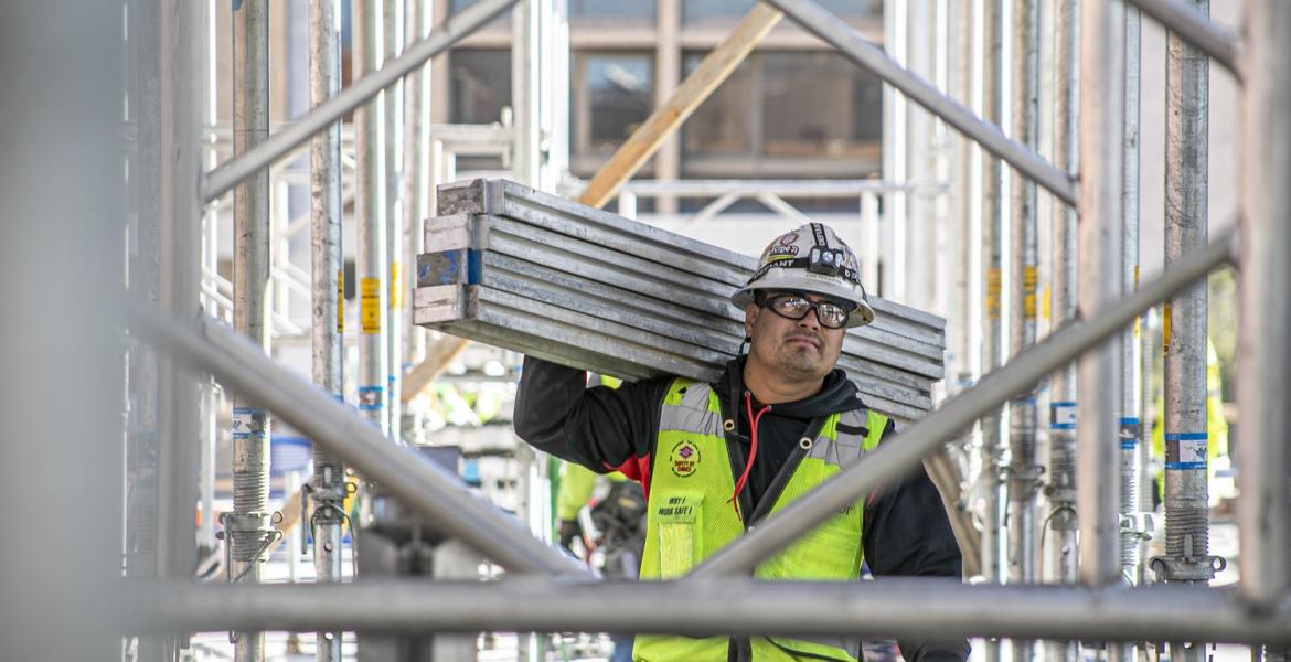 A man wearing a construction vest carries four metal beams on a construction site.