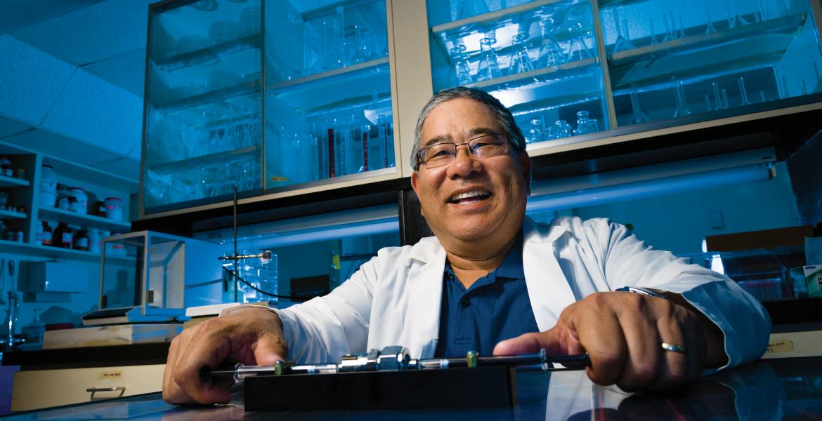 Terry Matsunaga in a laboratory lit with blue light, wearing a lab coat and smiling.