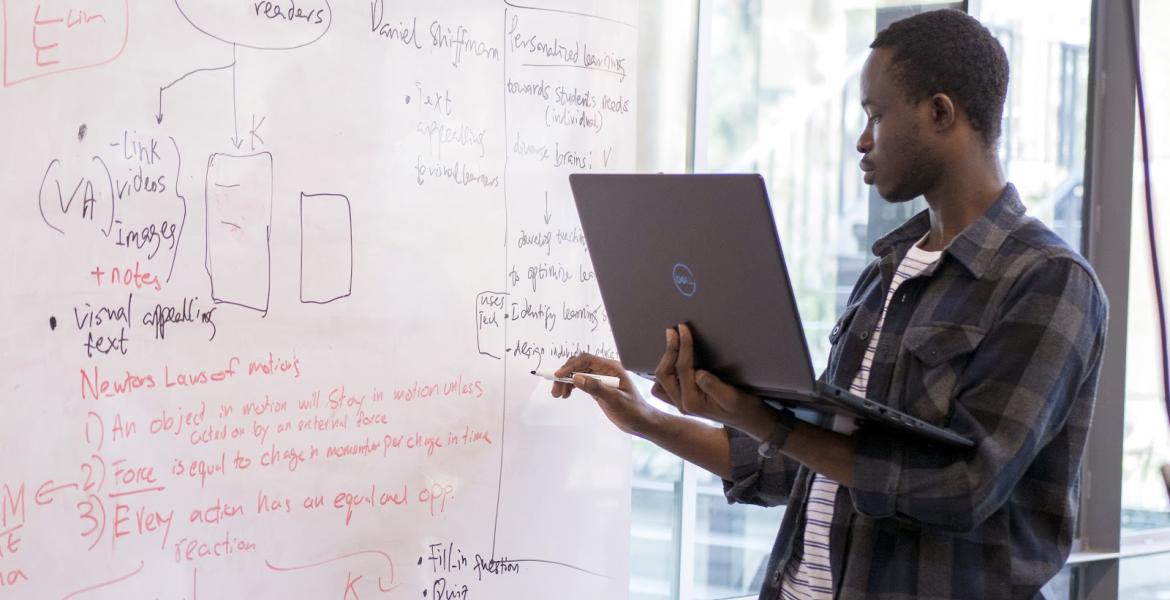 A student holds a laptop and writes on a dry erase board.