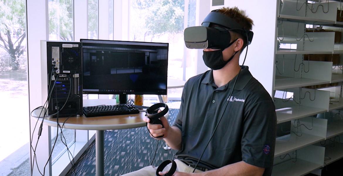 A student sitting next to a computer monitor and wearing a VR headset.