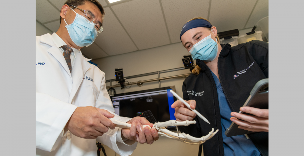 A professor and a student, both wearing surgical masks, seen from below. They are looking down at a model of the hand and wrist bones.