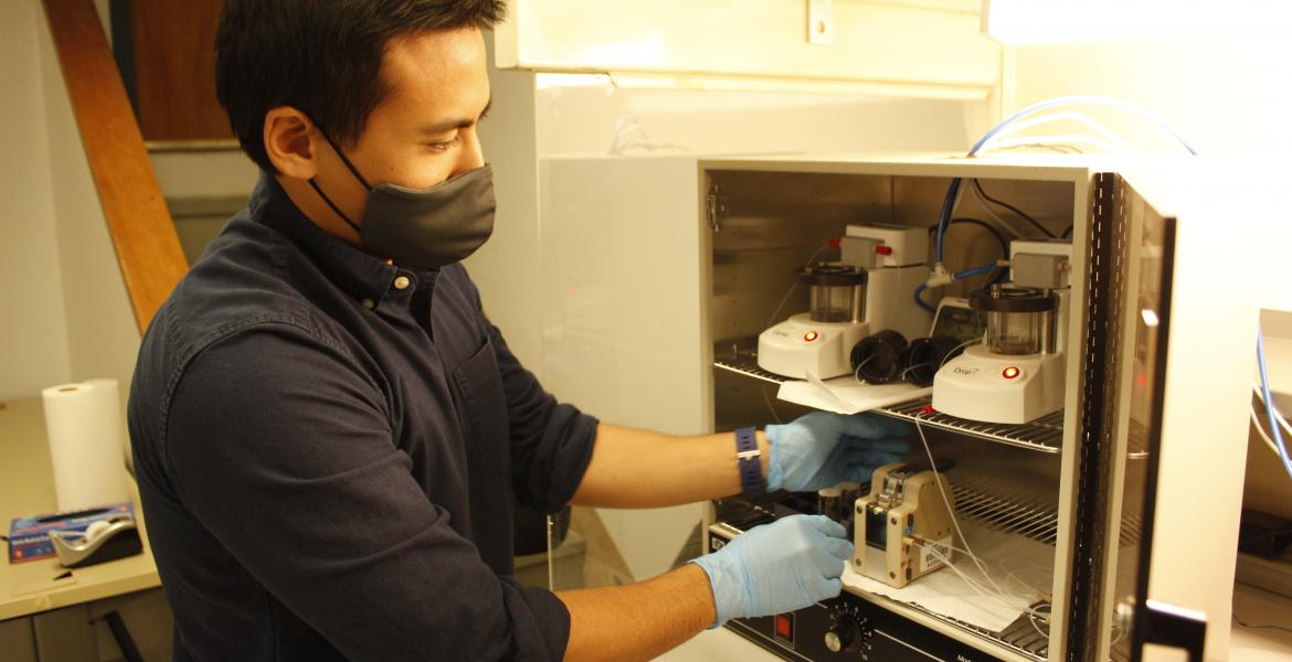 Suchol Savagatrup, wearing a face masks and blue latex gloves, reaches into a cabinet in his lab.