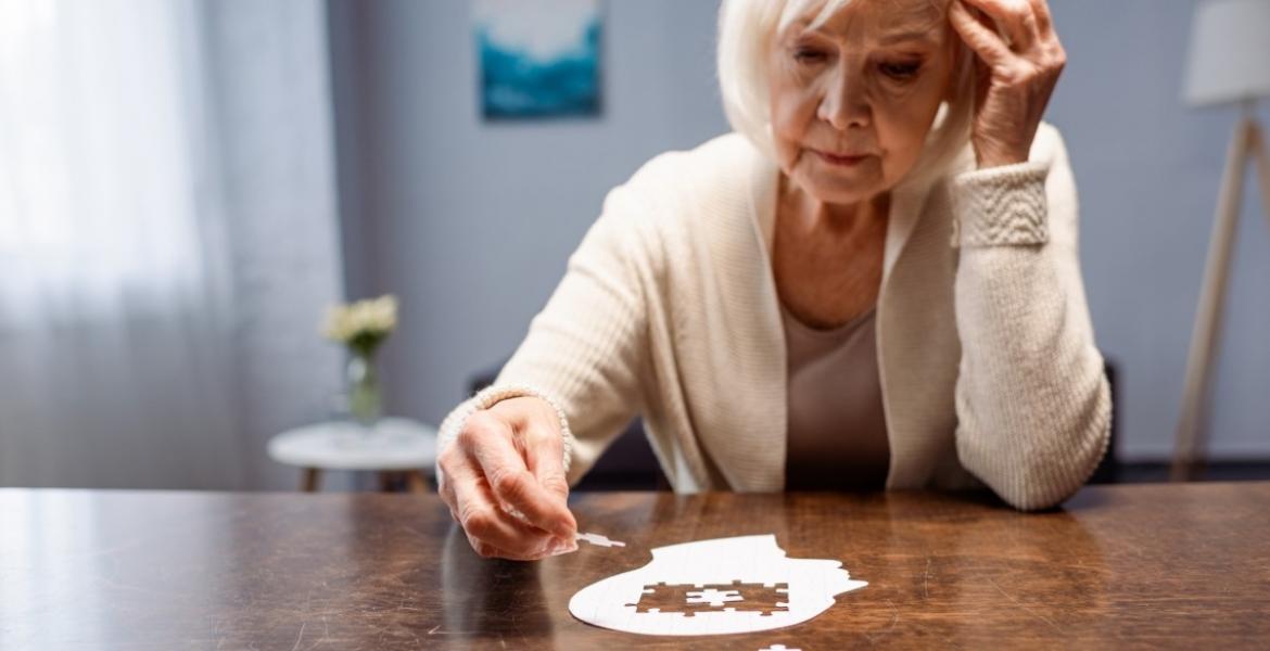 An older woman sitting at a table completing a white puzzle shaped like a human head.