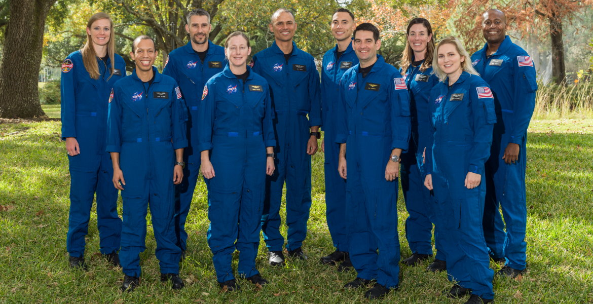 A group of ten astronauts stand in matching blue jumpsuits in a green outdoor area.