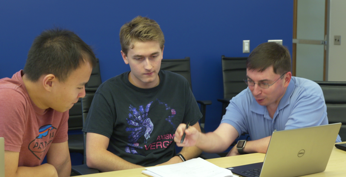 Three men lean over a laptop and set of papers on a table.