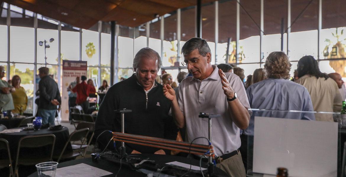 Two men stand in a crowded lobby. One is gesturing at machine on a table in front of him, while the other listens carefully.