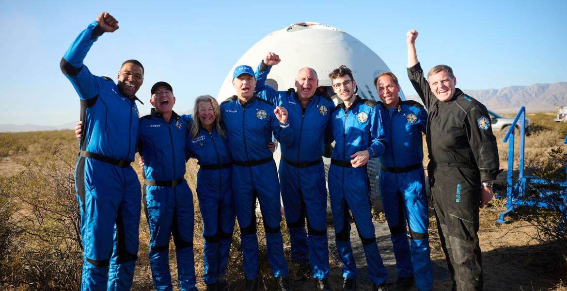 A group of eight people in blue jumpsuits smile in front of a round spacecraft. They look happy, with their arms around each other or raised in the air in celebration.