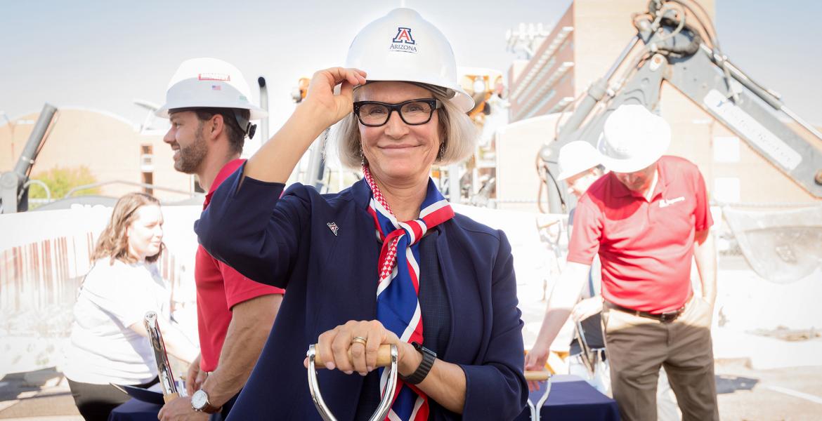 Betsy Cntwell on a construction site, wearing a white hard hat with a Block A logo. She is smiling and holding the bill of the hat.