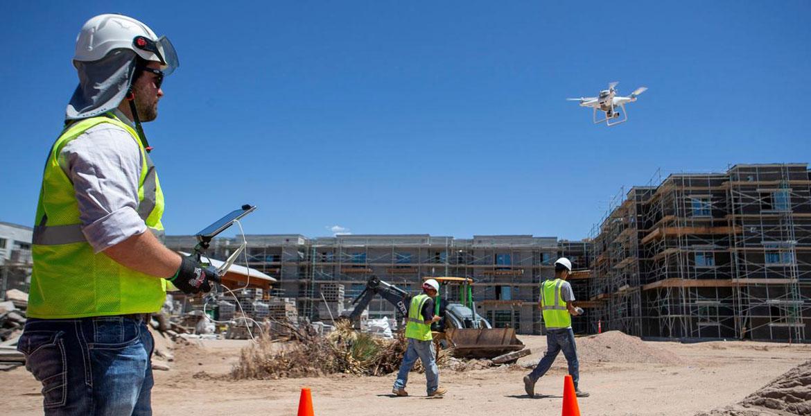 A man in a yellow construction vest and hard hat operates a drone on a construction site.