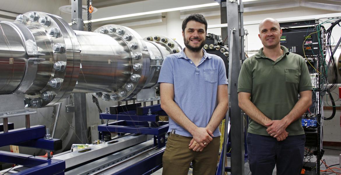 Alex Craig and Jesse Little standing next to a hypersonic wind tunnel, a horizontal metal tube about the circumference of a large tree.