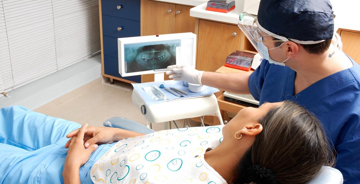 A dental patient lies in a chair while a dentist gestures toward X-rays of the patient's teeth.