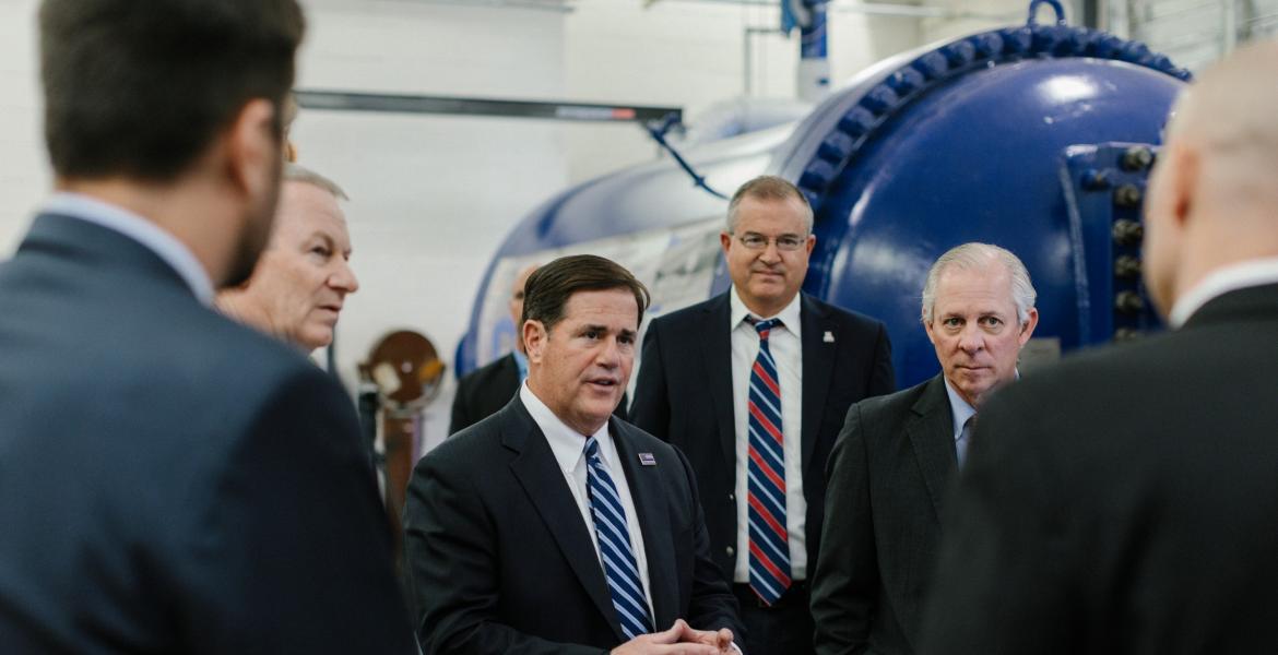 Six men stand next to a blue wind tunnel talking.