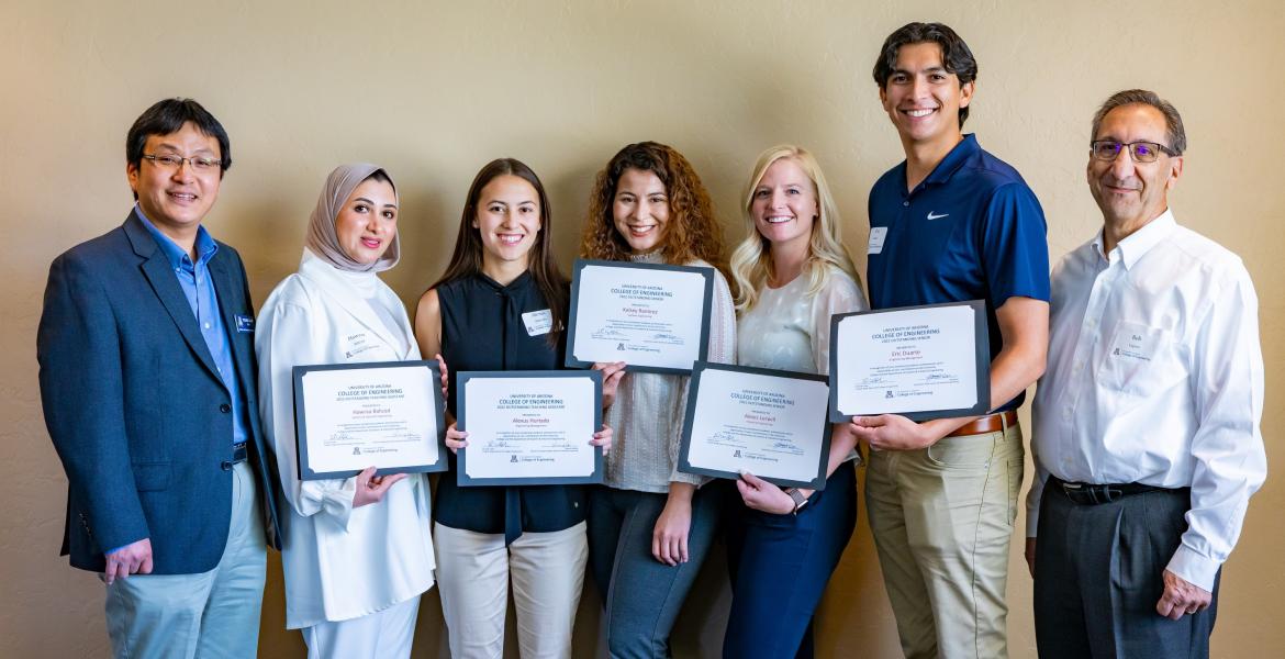 A group of seven people -- five students holding certificates flanked by two professors -- pose for a photo