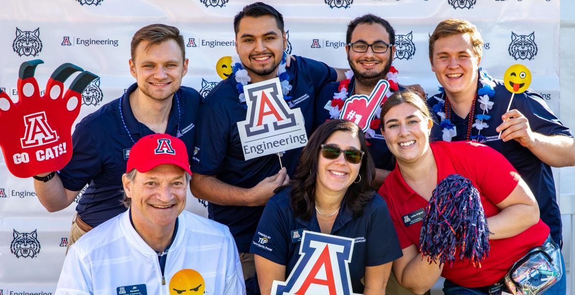 A group of seven people pose for a photo in front of a UA Engineering backdrop. They are smiling and holding props like a Block A and a foam finger in UA colors.