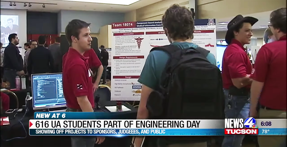 A student in a red polo stands in front of a scientific poster talking to someone in a green shirt reading the poster.