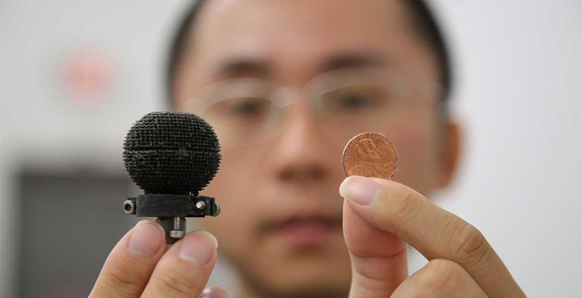 A man holds up a small 3D-printed black sphere in one hand, a penny in the other. The sphere is only about 3 times the size of the penny.