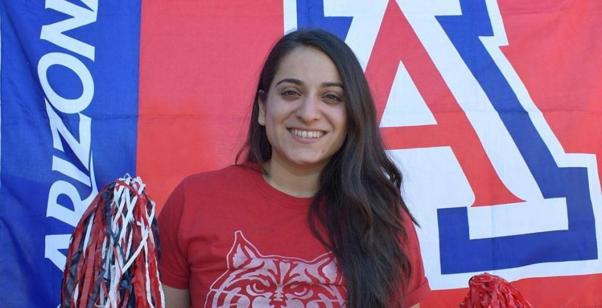 Maira Garcia smiles and holds two pom poms. Behind her is the University of Arizona Block A logo.