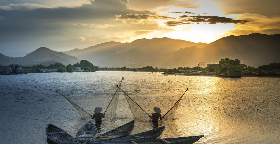 two fishers standing in water and holding up nets. There are four canoes behind them. In the distance is a sunset over mountains.
