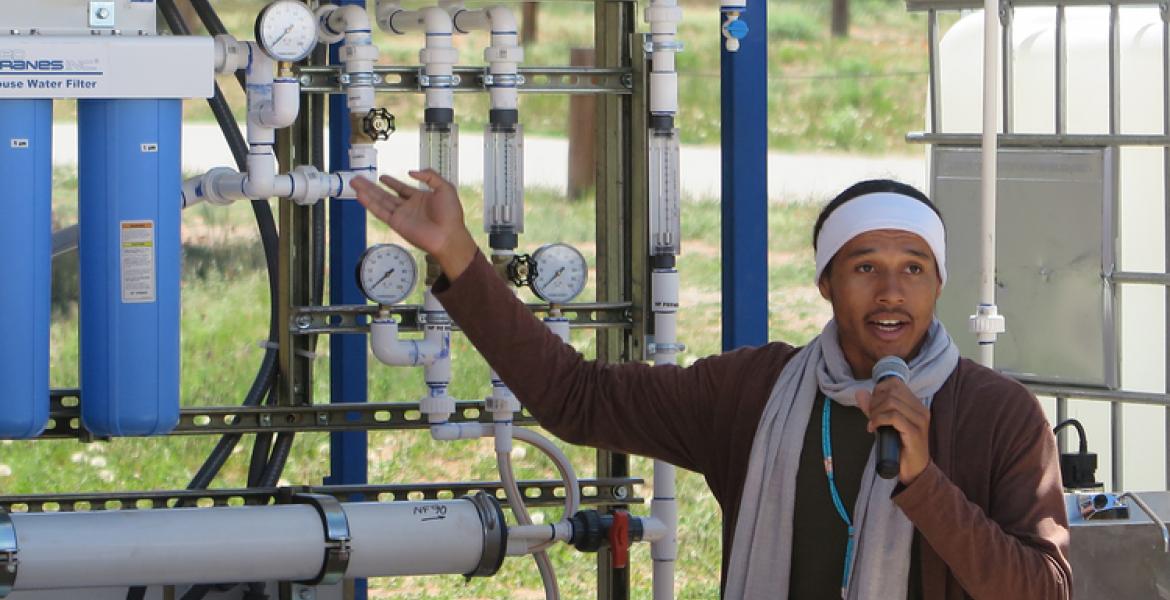 A young man holding a microphone gestures toward a systems of pipes and gauges.