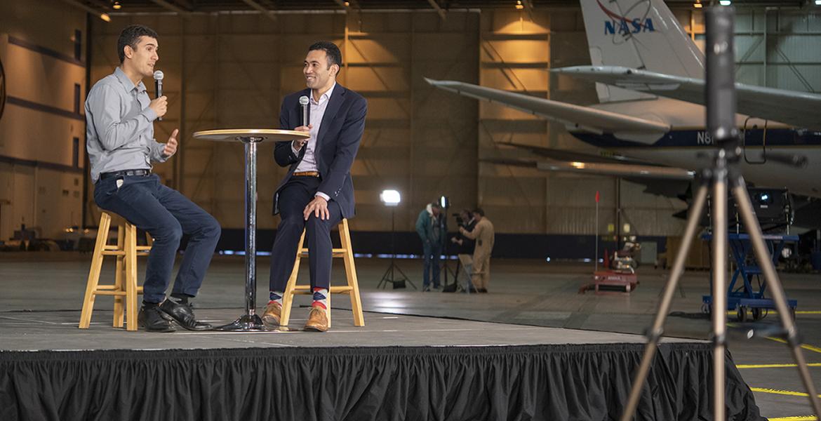 Two men sit on a stage with a NASA plane in the background